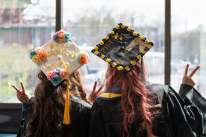 Two women looking out a window with their backs facing the camera, wearing graduation robes and decorated caps, giving the peace sign
