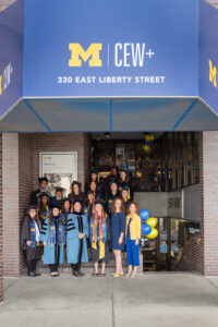 Group of CEW+ scholars in graduation robes posed together on the front stairs of CEW+ with the maize and blue CEW+ awning above them