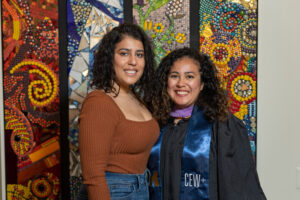 Two women of undetermined race smiling and embracing in front of multicolored mosaic wall art