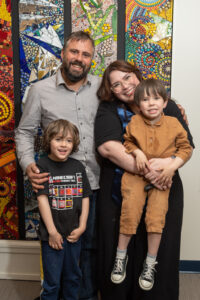 Caucasian family with man, woman and two children embracing in front of multicolored mosaic wall art