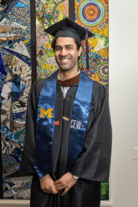 Man of undetermined ethnicity wearing graduation robe and cap smiling in front of multicolored mosaic wall art