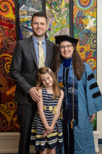 Caucasian family with one man, one woman wearing graduation robe and cap, and one daughter smiling in front of multicolored mosaic wall art