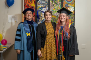 Three women smiling - one woman of undetermined ethnicity, one Caucasian woman wearing a graduation robe, CEW+ stole, graduation cap, and one multiracial woman wearing a graduation robe, CEW+ stole, graduation cap smiling in front of multicolored mosaic wall art