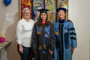 Three women smiling, one woman of undetermined ethnicity wearing a graduation robe, CEW+ stole, graduation cap, one Caucasian woman, and one multiracial woman wearing a graduation robe, CEW+ stole, graduation cap smiling in front of multicolored mosaic wall art