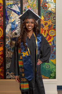 Black woman wearing graduation robe and cap smiling in front of multicolored mosaic wall art