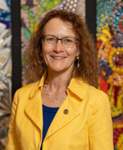 Doreen Murasky wearing a yellow blazer and blue shirt standing in front of colorful mural